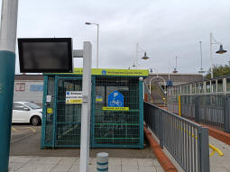 Facing away from the end of the tram line with the railway platform on the right, a hard-to-read-during-daylight tram and (I think?) bus departure display stands in front of a cycle park